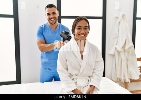 Latino uomo e donna che indossano la fisioterapia uniforme massaggiante collo con pistola a percussione al centro di bellezza Foto Stock