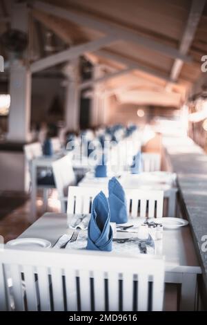 Un focus selettivo su un tavolo da pranzo di legno bianco, argenteria, e tovaglioli blu in un ristorante su un portico di un resort tropicale nel pa Foto Stock