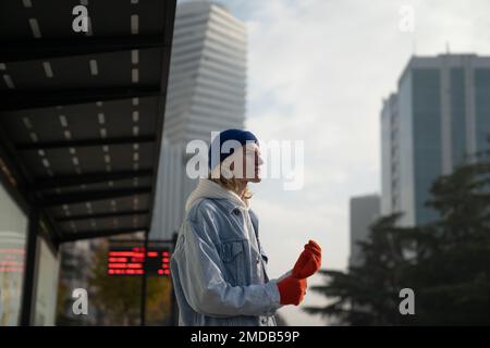 Giovane ragazzo di tendenza Hipster in piedi alla fermata dell'autobus cittadino sullo sfondo urbano Foto Stock