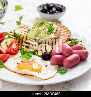 Uovo fritto con mini salsiccia, pomodori, zucchine alla griglia, olive nere e toast con guacamole. Colazione all'inglese. Brunch. Foto Stock