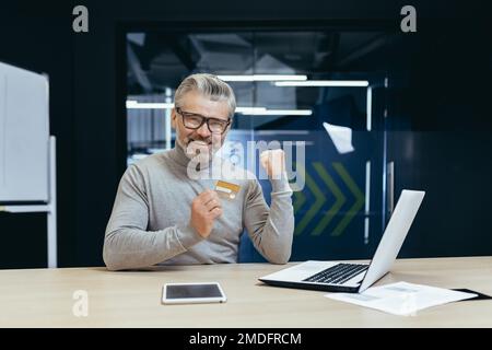 Il riuscito uomo d'affari grigio haired gioisce e celebra il completamento della transazione di trasferimento di denaro, ritratto del capo all'interno dell'ufficio con la carta di credito della banca e il laptop, uomo maturo che guarda la macchina fotografica. Foto Stock