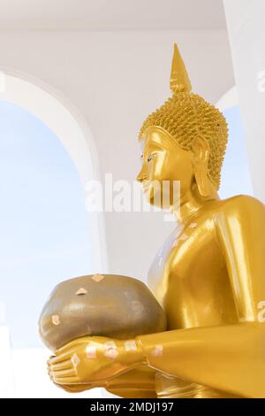 Un Buddha d'oro che regge una statua di una ciotola di elemosina in un antico stupa contro un arco bianco e un cielo blu sullo sfondo. Thailandia. Foto Stock