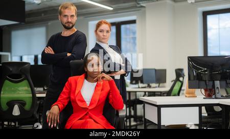 Donna caucasica dai capelli rossi, uomo caucasico bearded in piedi dietro una giovane donna afroamericana seduta in ufficio. Foto Stock