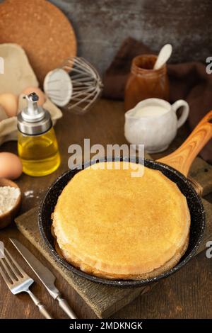 Festeggiamo la giornata delle fritture, cucinando una sana colazione. Deliziose crepes o frittelle fatte in casa in una padella e ingredienti su un tavolo rustico. Foto Stock