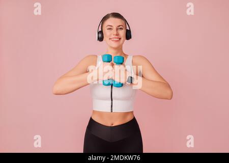 Ritratto di felice signora caucasica in abbigliamento sportivo con manubri blu su sfondo rosa. Giovane donna allegra con cuffie che indossano abbigliamento sportivo Foto Stock