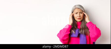 Primo piano di moda donna anziana sensazione di malessere, toccando la testa e grimacing da mal di testa, lamentarsi di emicrania, sfondo bianco Foto Stock