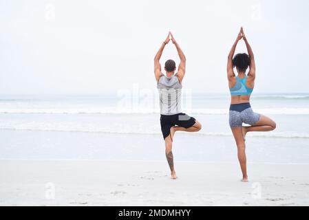 L'equilibrio è che cosa conduce all'adempimento. Ripresa retroversa di un giovane uomo e di una donna che praticano lo yoga insieme in spiaggia. Foto Stock