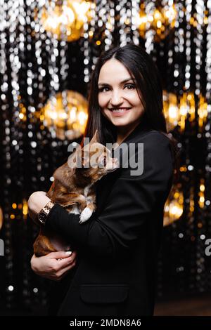 Giovane bella bruna sorridente femmina in giacca nera tenendo il suo cane cucciolata nelle sue mani. Donna spensierata che posa vicino a una parete di tinsel lucido in studio wi Foto Stock