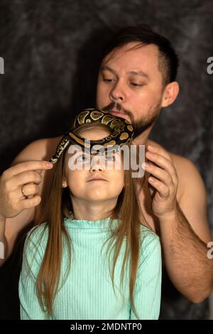 Padre e figlia giocano con serpente. Ritratto di ragazza sorridente in verde e uomo che tiene serpente su sfondo grigio. Messa a fuoco selettiva. Foto di alta qualità Foto Stock