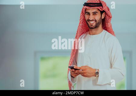 Giovani uomini d'affari arabi musulmani moderni che indossano abiti tradizionali mentre usano gli smartphone a casa. Foto Stock