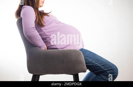 Ragazza incinta si siede su una sedia sul posto di lavoro, sfondo bianco. Il concetto di congedo di maternità e le condizioni di lavoro ai sensi della legge. Vantaggi e ma Foto Stock