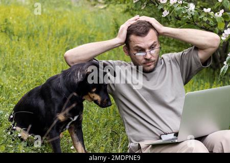 Uomo e cane che guardano lo schermo del notebook con emozioni stupite hanno scioccato l'espressione facciale nelle mani mentre si siedono sull'erba verde all'aperto. Foto Stock