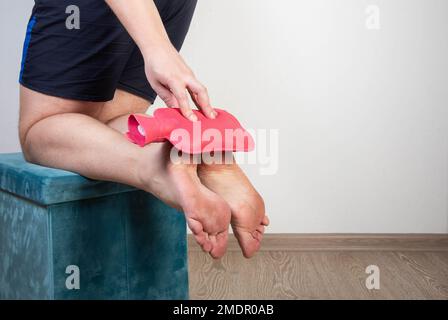 Un uomo tiene un cuscino riscaldante con acqua calda vicino alle suole dei suoi piedi. Rilievo di dolore e trattamento per il dolore dello sperone del tallone. Foto Stock
