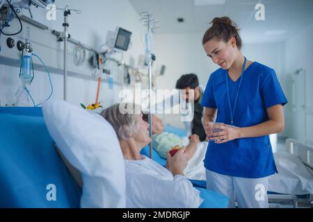 Giovane infermiera che dona un bicchiere d'acqua al paziente anziano del reparto ospedaliero. Foto Stock