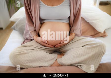 Donna incinta felice che le accarezzava il ventre, seduta a letto. Foto Stock