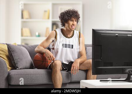 Un ragazzo in una maglia sportiva con un baskteball seduto su un divano a casa e guardando la tv Foto Stock