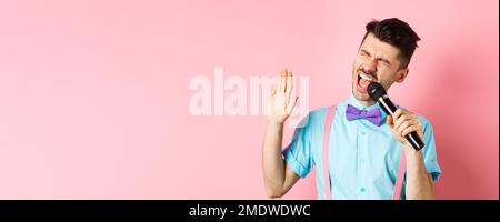 Concetto di festa ed eventi di festa. Il ragazzo divertente esegue una canzone, cantando il karaoke in microfono con la faccia spensierata, in piedi in bow-craving e sospenders sopra Foto Stock