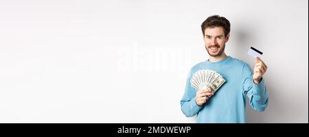 Bel giovane uomo sorridente e che offre il pagamento in contanti e senza contatto, mostrando denaro con carta di credito in plastica, in piedi su sfondo bianco Foto Stock