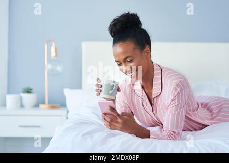 Tempo di recuperare un po' di pettegolezzi caldi. una giovane donna attraente che beve caffè mentre usa il suo cellulare a letto a casa. Foto Stock