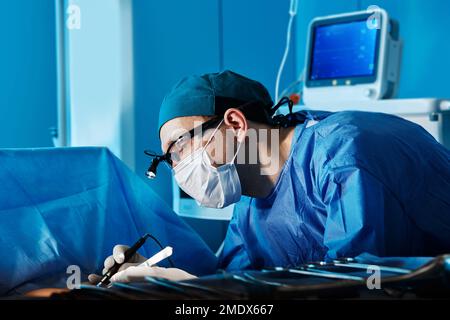 Ritratto del chirurgo focalizzato indossando maschera protettiva e proiettore durante il funzionamento nella sala operatoria dell'ospedale. Professione di chirurgo Foto Stock