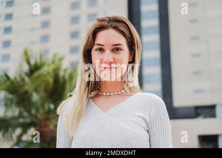 Primo piano ritratto di una felice bionda donna sorridente guardando la macchina fotografica e toccando i suoi capelli. Vista frontale di giovane donna isolata con denti perfetti che ridono all'aperto. Foto di alta qualità Foto Stock