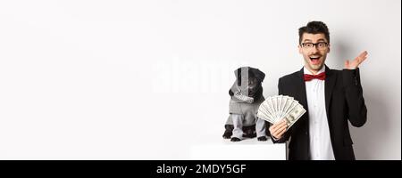Felice giovane uomo e carino cane nero in piedi in costumi di festa, proprietario di pug che tiene i soldi dollari, fissando la macchina fotografica stupito, sfondo bianco Foto Stock