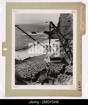 Moderni Cliff-abitanti (7 di 7). Sulla piattaforma mostrata qui, a 150 piedi sopra la stretta spiaggia rocciosa, si trovano il derrick, la casa del derrick e i tamburi d'olio alla Stazione leggera di Cape Hinchinbrook, Alaska. Oltre, le scogliere che si innalzano si aggirano a 1.400 piedi direttamente dal mare. I guardiani costieri a piedi nudi danno un riposo ai muscoli stanchi dopo che i tamburi sono stati riempiti con un anno di rifornimento di olio combustibile portato alla stazione dalla Guardia Costiera Icebreaker NORTHWIND. Gli uomini hanno rimosso l'ingranaggio del piede imbevuto da acqua di mare fredda durante l'operazione. Nelle prime fasi del lavoro di due giorni, il tempo 'chiuso', marooning 22 offic Foto Stock