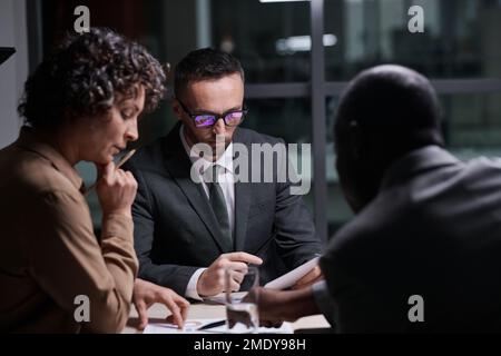 Concentratevi sull'analisi di un documento finanziario da parte di un broker serio di fronte a due colleghi interculturali che discutono di dati grafici durante la riunione Foto Stock