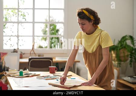 Giovane artigiano ispirato in casualwear in piedi da tavolo con forniture di disegno e la creazione di nuove opere d'arte o schizzo su carta in studio Foto Stock