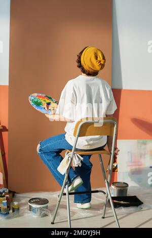 Vista posteriore della giovane pittura creativa femminile che tiene la tavolozza con pitture acriliche di vari colori mentre lavora su un nuovo capolavoro Foto Stock