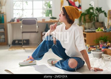 Giovane donna penitente in casualwear tenendo la matita per la bocca mentre si siede sul pavimento con le carte e pensando a nuove opere d'arte Foto Stock