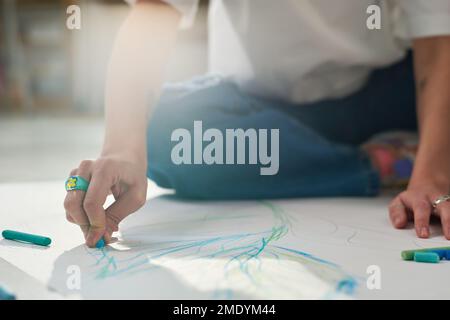 Primo piano delle mani di una giovane artista creativa con pastelli blu su carta bianca mentre si siede sul pavimento dello studio di casa Foto Stock