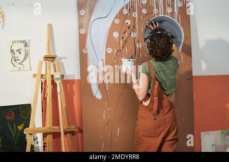 Vista posteriore della giovane donna creativa con pitture che creano nuove opere d'arte astratte mentre si trova davanti al pannello nel suo studio domestico Foto Stock