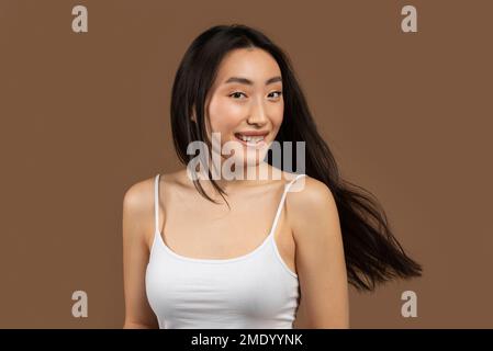 Ritratto di felice donna coreana posa con lunghi e sani capelli volanti su sfondo marrone e sorridente alla macchina fotografica Foto Stock