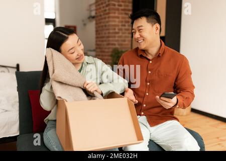 Shopping online di successo. Felice cliente asiatico femmina disimballaggio consegnato scatola dal negozio, seduto con husbnad sul divano Foto Stock