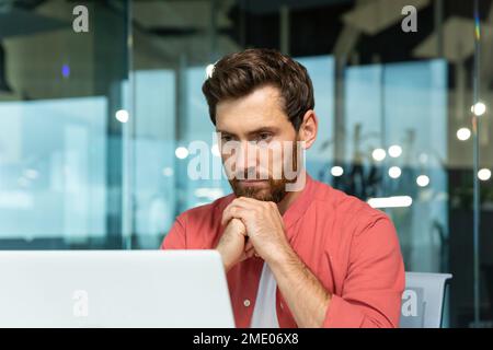 Serio uomo d'affari pensante primo piano, uomo in camicia rossa casual che lavora all'interno di un ufficio moderno, capo maturo che lavora con il portatile sul posto di lavoro mentre si siede Foto Stock