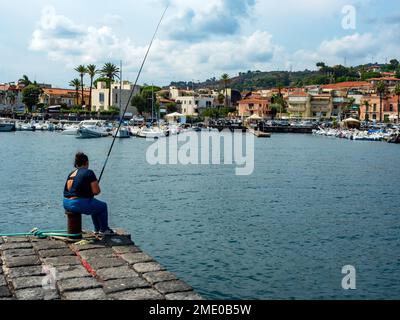 Acitrezza Sicilia Italia 10-02-2021: Vista da dietro, giovane donna seduta su un molo intenta a pescare nel porticciolo di un caratteristico villaggio di pescatori Foto Stock