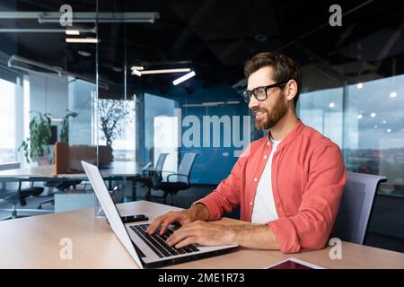 Uomo sorridente di successo che lavora all'interno dell'ufficio con il laptop, uomo d'affari in camicia rossa sorridente e che digita sulla tastiera negli occhiali, programmatore che lavora software per il programma. Foto Stock