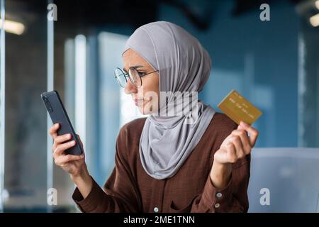 Primo piano. Una giovane donna araba in un hijab e occhiali sta tenendo un telefono e una carta di credito. Shopping online, pagamento, conto corrente. Foto Stock