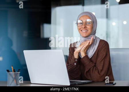 Ritratto di una giovane bella studentessa araba in hijab. Seduto al tavolo in ufficio con un computer portatile, guardando la fotocamera, sorridendo, ripiegò le mani insieme. Foto Stock