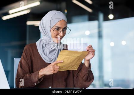 Una giovane studentessa araba felice in hijab. Seduto alla scrivania in ufficio, con una busta con una lettera, ha ricevuto una notifica di ammissione all'università, i risultati degli esami. Foto Stock