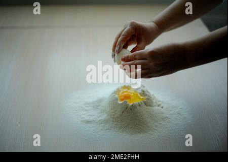 mani femminili primo piano rompere uovo di pollo bianco e versare tuorlo e proteine nel mucchio di farina piccole gocce raggiungere per mano scavare su tuorlo uovo lentamente scorre verso gocce con caduta Foto Stock