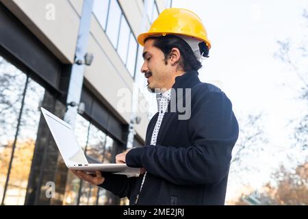 Dal basso vista laterale di un giovane ingegnere maschile ispanico positivo in abiti casual intelligenti e casco giallo che lavora sul computer portatile mentre si trova vicino al moderno b Foto Stock