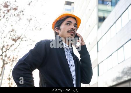 Da sotto di concentrato giovane imprenditore etnico maschile con capelli scuri e barba in abiti casual elegante e hardHat parlare su smartphone mentre si è in piedi Foto Stock