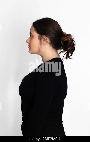 Vista laterale di una giovane donna seria con un'acconciatura mordente in maglione nero su sfondo bianco Foto Stock