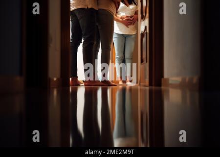Ritagliare una coppia irriconoscibile in abiti casual in piedi di fronte allo specchio nel corridoio scuro e l'uomo che abbraccia la donna che trasporta il bambino da dietro Foto Stock