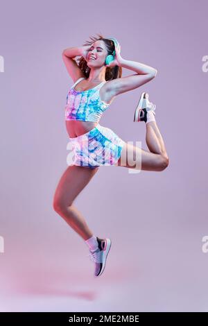 Un'atleta femminile completamente in forma con una gamba sportiva colorata che alza mentre salta in uno studio toccando le cuffie ascoltando musica danzante Foto Stock