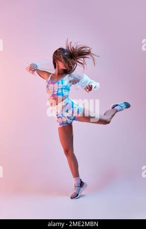 Giovane donna snella in abbigliamento sportivo stretto che alza le braccia e le gambe mentre ballano sullo sfondo rosa in studio Foto Stock