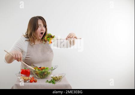 divertente donna allegra mangiare insalata su sfondo bianco si apre bocca larga rigonfiamenti occhi grande cucchiaio di legno stuffs porzione di cibo sulla tavola ingredienti spazio per testo perdita di peso annuncio Foto Stock