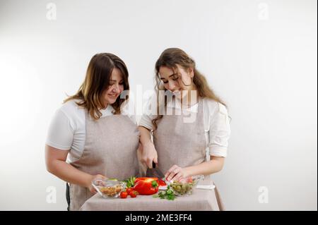 Donne madre e figlia preparare insalata mamma assomiglia a 16-18 anni figlia tagli rosso peperone ingredienti identici grembiuli cucina bianco spazio di sfondo per l'insegnamento del testo scuola di cucina Foto Stock
